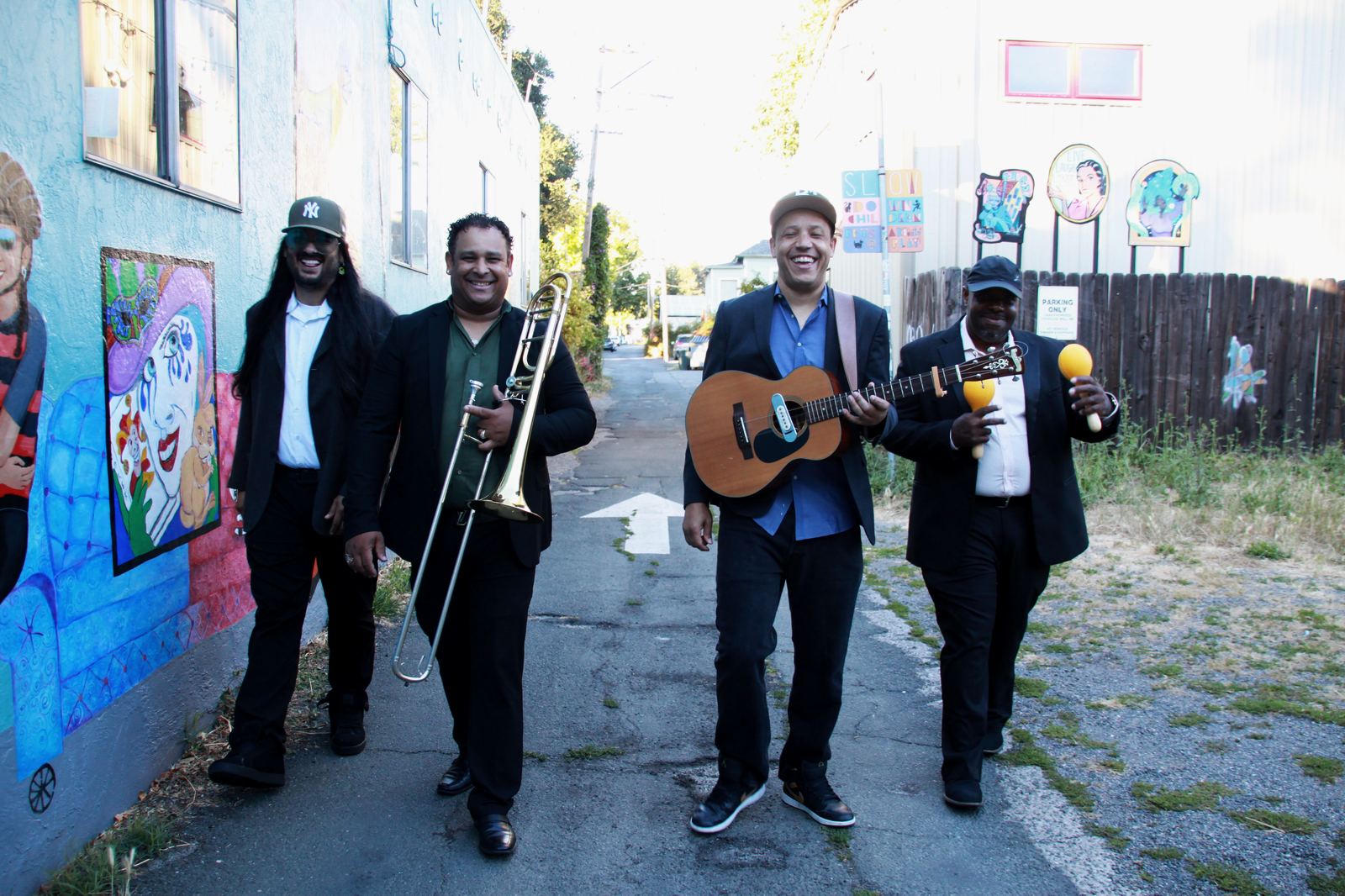 Five members of Son Chévere walking up a sunny alley past a painted mural, carrying a trombone, a tres cubano and maracas.