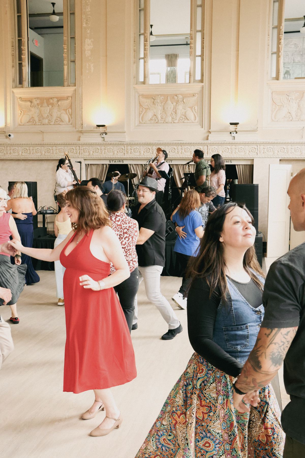 Couples dancing on a ballroom floor, a woman in a red dress in front, with Son Chévere on stage behind them.