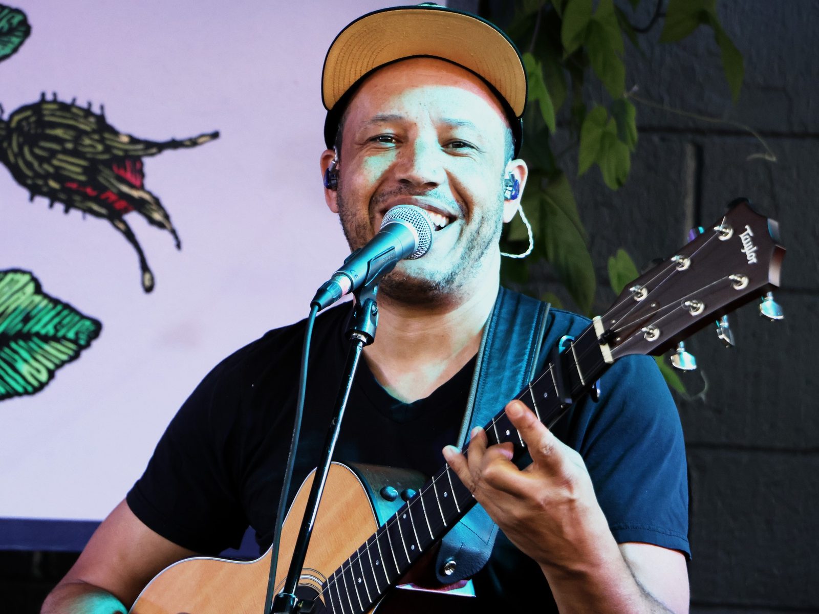 Jordan Wilson grinning at the microphone as he plays his tres cubano, in front of a painted mural.