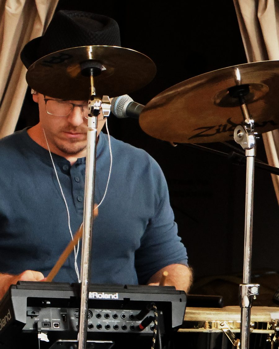 Ryan Mead in a black hat and glasses, playing sticks on his percussion pad behind the cymbals.